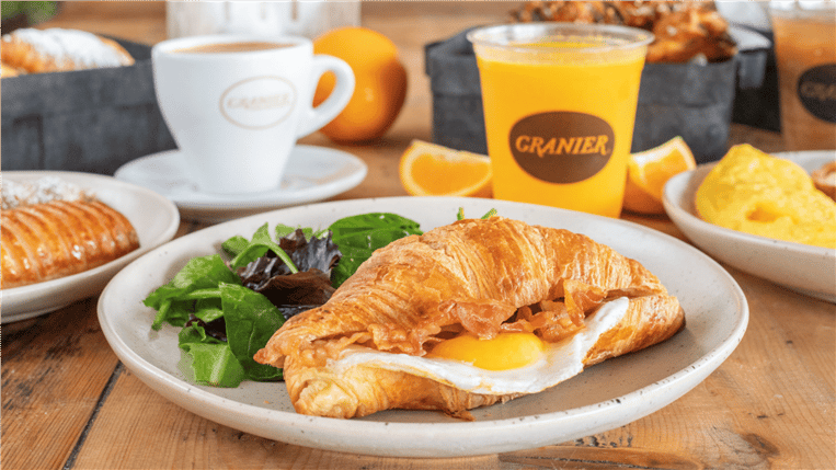 Croissant sandwich with egg and bacon. Assorted other breakfast items around it on a wood table.
