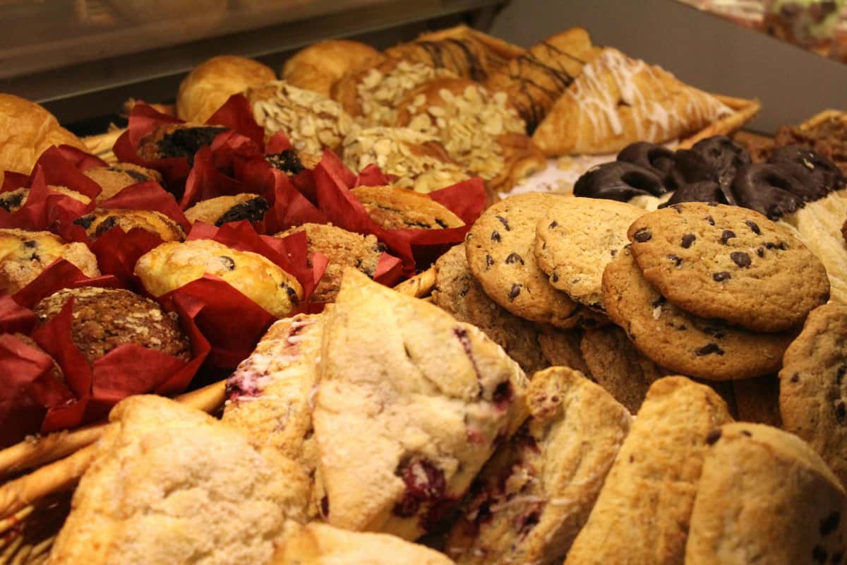 A variety of freshly baked pastries, cookies, and muffins arranged in red paper wrappers, including scones, croissants, and chocolate chip cookies.