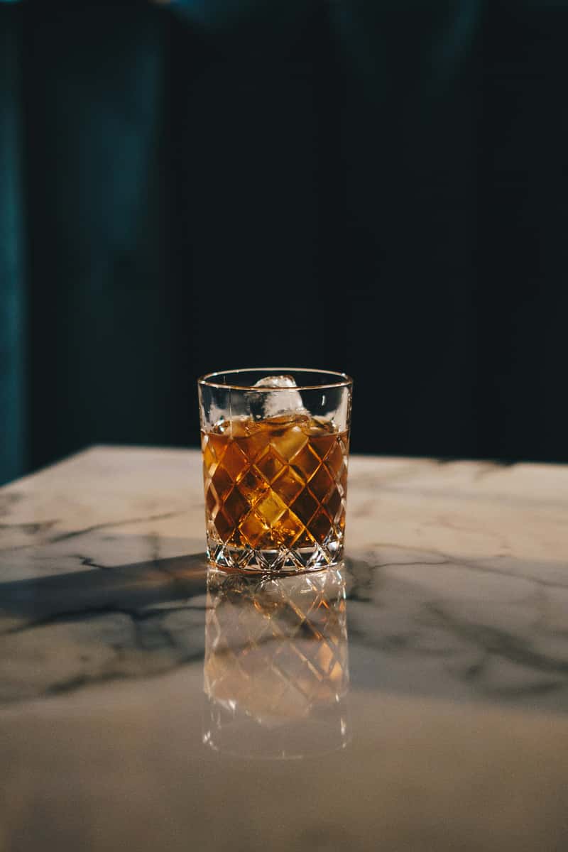 Whiskey cocktail with ice in a cut-glass tumbler on a polished marble table, reflecting a sophisticated bar atmosphere with dark velvet seating.