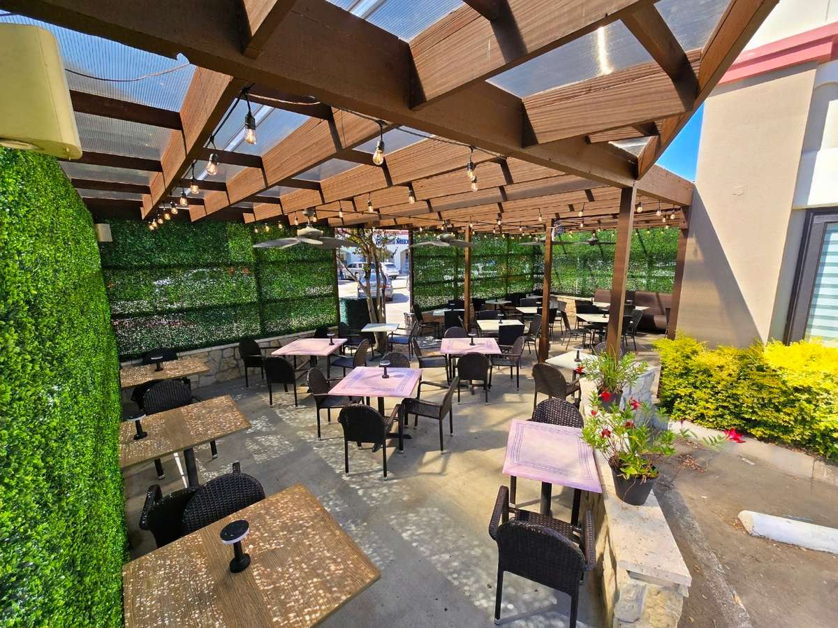 An outdoor patio dining area with multiple tables and woven chairs arranged under a wooden pergola. String lights hang overhead, and green hedge walls surround the space, creating a cozy atmosphere. The patio is decorated with potted plants and receives bright natural sunlight.