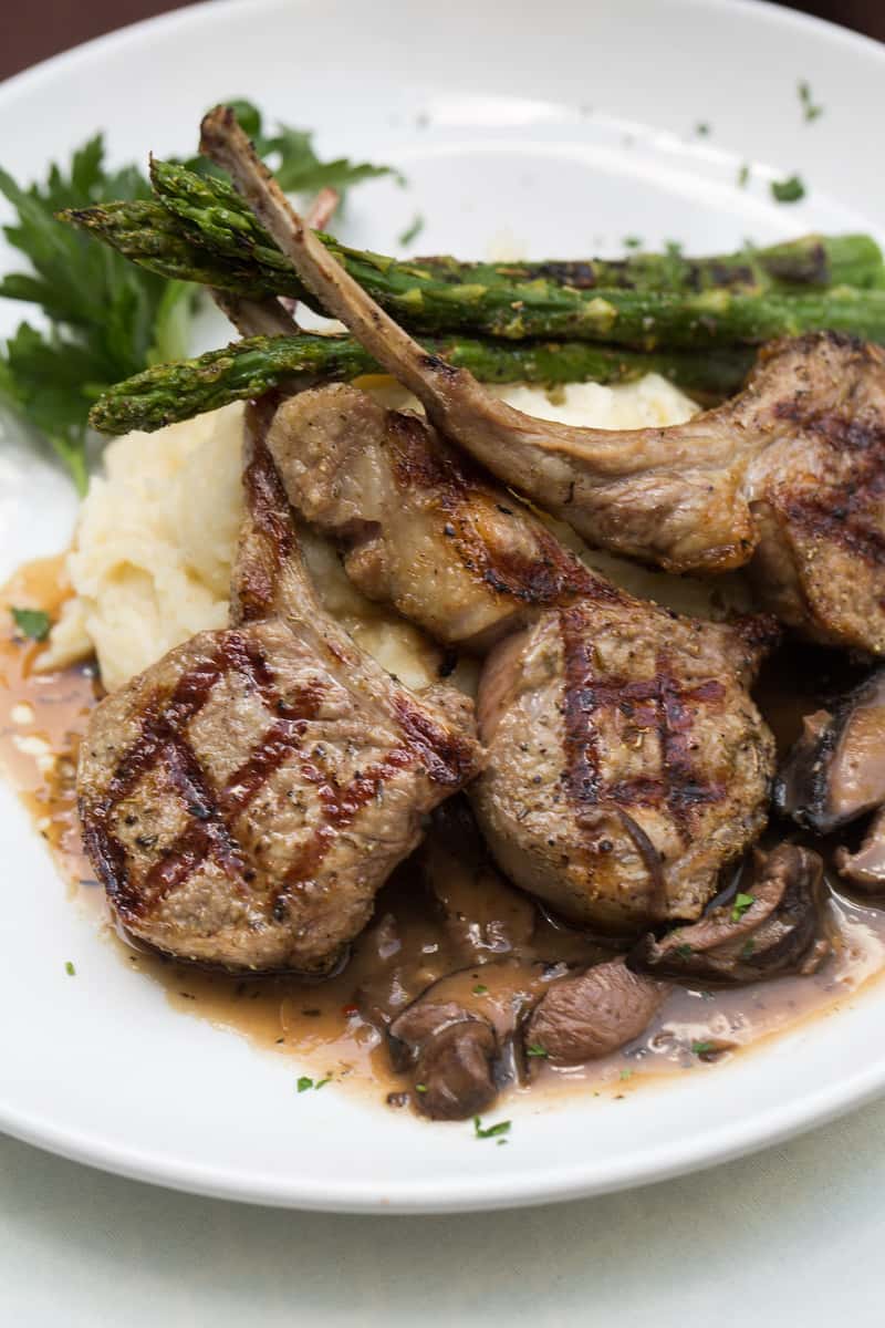 Grilled lamb chops served over mashed potatoes with sautéed mushrooms and sauce, topped with grilled asparagus spears and garnished with fresh parsley.
