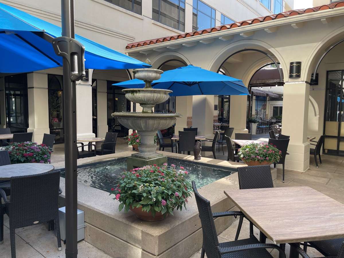 Outdoor courtyard restaurant patio with a central stone fountain, blue umbrellas, and surrounding tables and chairs in a Mediterranean-style setting.