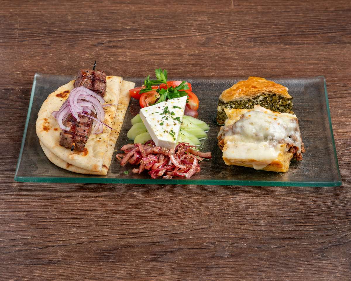 A Mediterranean-style sampler plate featuring a grilled beef skewer wrapped in pita with sliced red onions, a wedge of feta cheese with cucumbers and cherry tomatoes, marinated red onions, a slice of spinach pie (spanakopita), and a portion of moussaka topped with melted béchamel, all served on a rectangular glass plate.