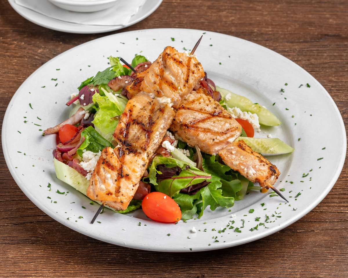 Grilled salmon skewers served over a fresh Greek-style salad with mixed greens, cherry tomatoes, cucumbers, red onions, and crumbled feta on a white plate.