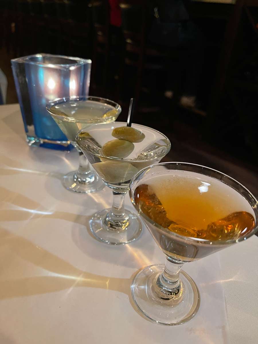 Three martinis lined up on a white tablecloth—one amber-colored, one classic with olives, and one pale—set beside a glowing blue candle holder in a dimly lit restaurant.