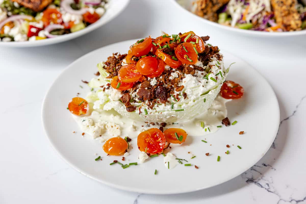 Jonathan the rub's classic wedge salad, topped with bright orange cherry tomatoes, crispy bacon bits, blue cheese crumbles, and creamy dressing on a white plate.