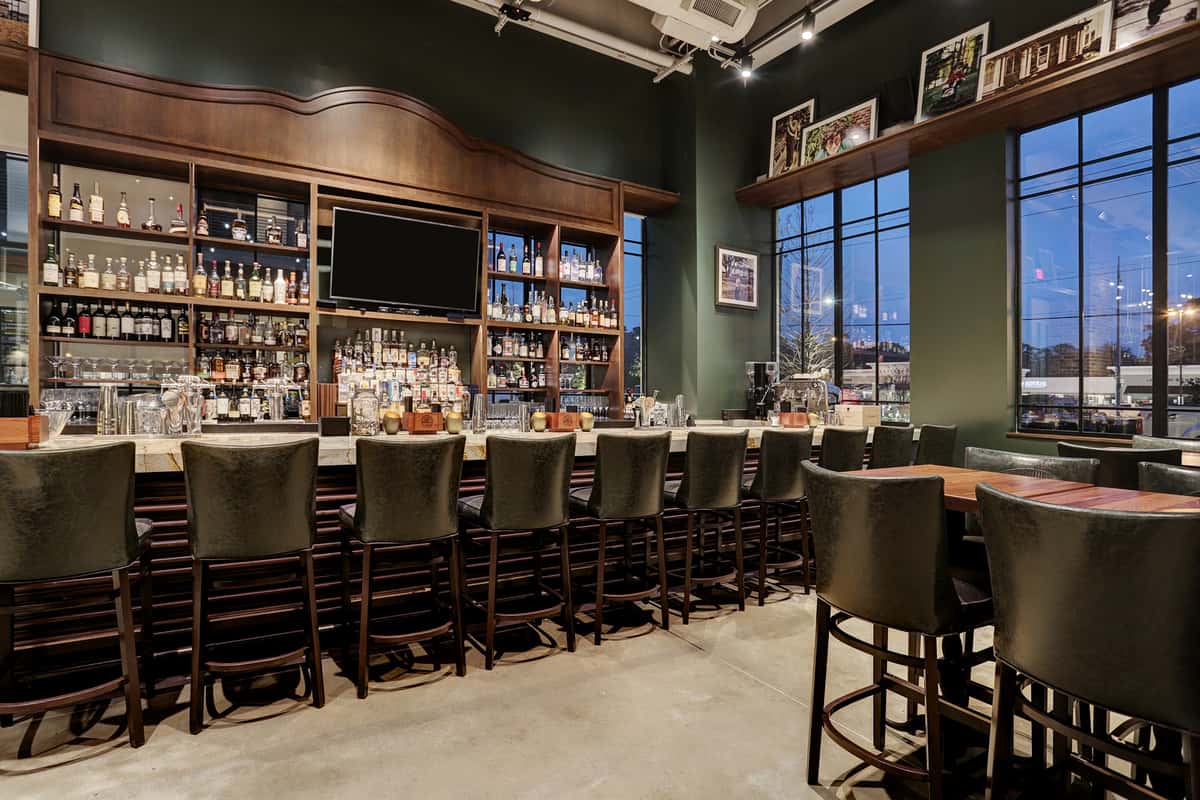Jonathan the rub restaurant bar interior with dark green walls, rich wood shelving, a marble countertop, and a row of dark leather bar stools.