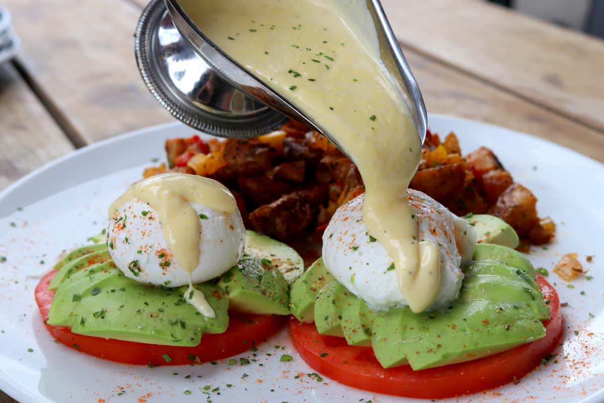 Hollandaise sauce poured over poached eggs, fresh green avocado, and red tomato slices, served with potatoes on a white plate.
