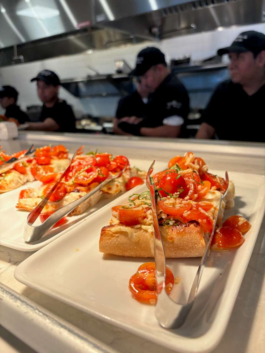 Bright red tomato bruschetta on white plates with serving tongs, set against a blurred background of chefs in black uniforms working in a stainless steel professional kitchen.