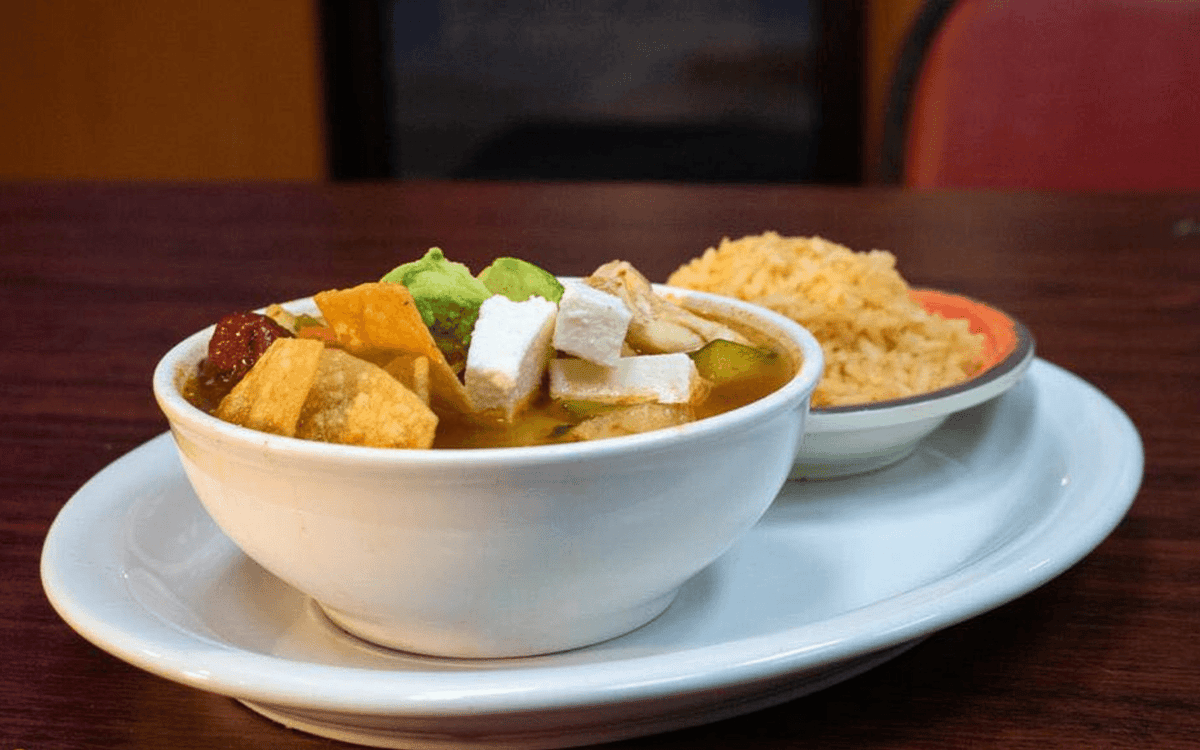 Authentic Mexican tortilla soup served in a white bowl, topped with crispy tortilla strips, avocado slices, queso fresco, and a side of Mexican rice. Perfect comfort dish at a traditional taqueria.