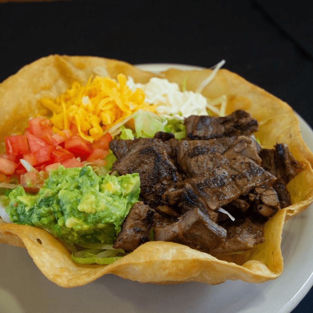 Taco salad served in a crispy tortilla bowl, filled with grilled steak, shredded lettuce, guacamole, diced tomatoes, shredded cheese, and sour cream — a hearty Mexican-style entrée.