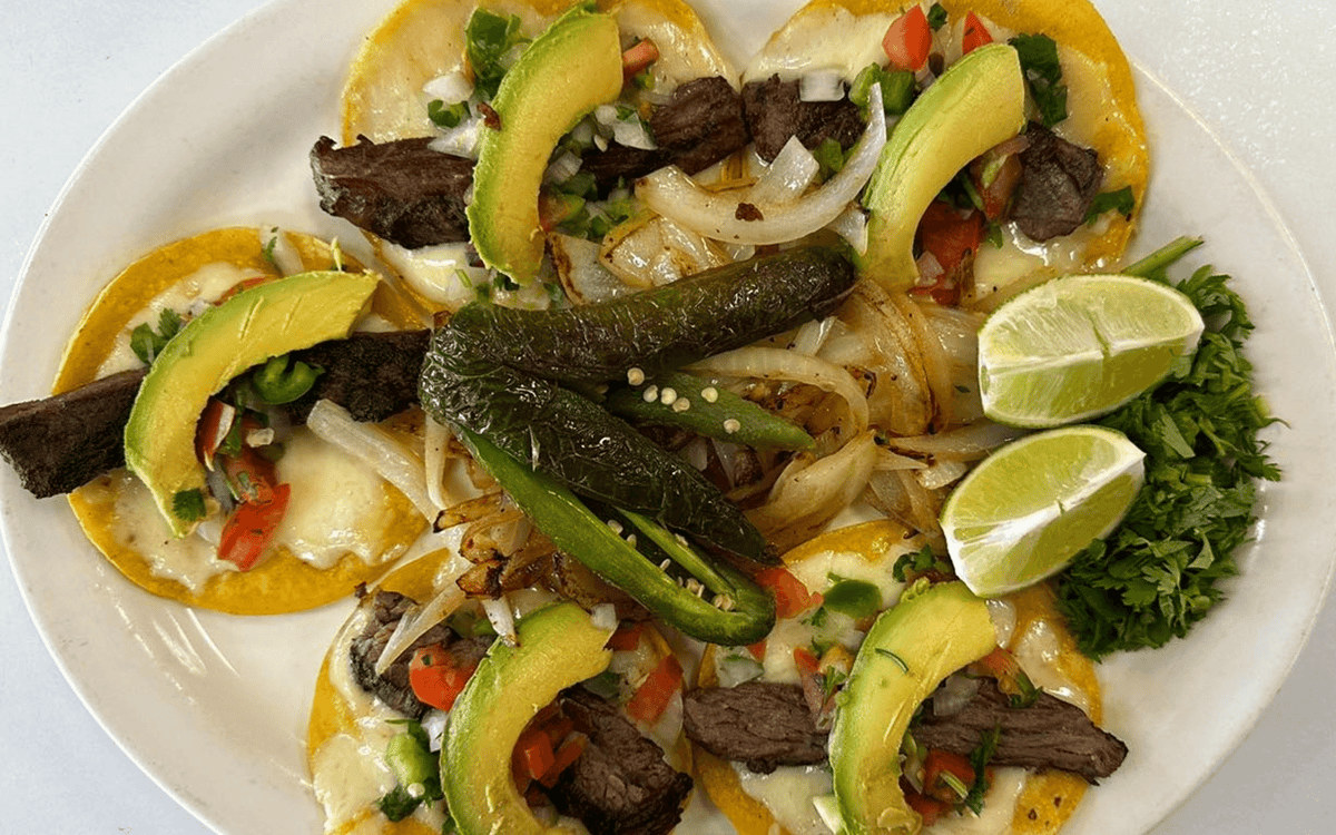 Plate of mini carne asada street tacos topped with grilled steak, avocado slices, sautéed onions, jalapeños, fresh cilantro, and lime wedges, served on a white dish — vibrant and authentic Mexican presentation.