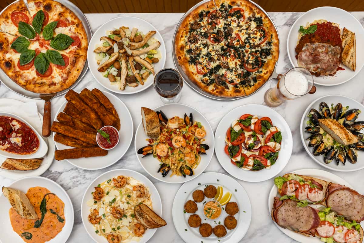 top-down spread of assorted pizzas, pasta, and appetizers arranged on a table for a family-style meal