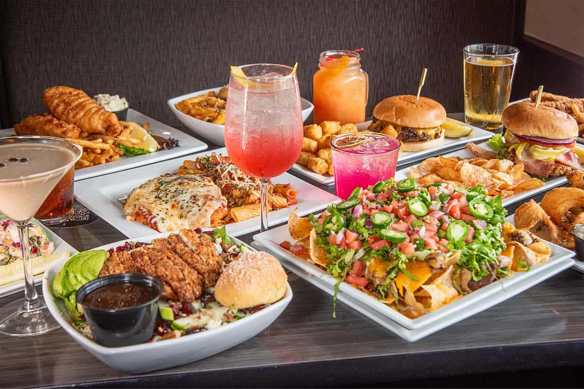 A full table featuring a variety of American dishes, including burgers, nachos, pasta, fried chicken, fries, tater tots, and colorful cocktails