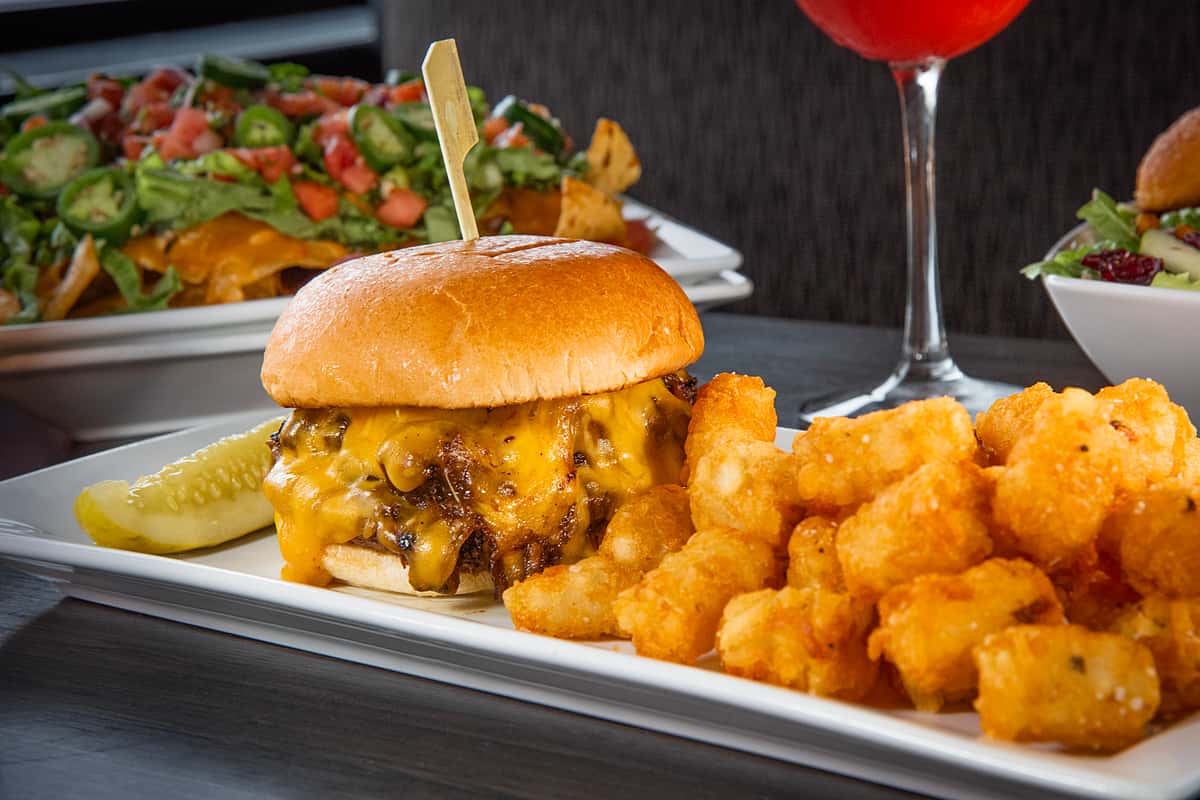 Cheeseburger with melted cheddar served with crispy tater tots and a pickle on a white rectangular plate