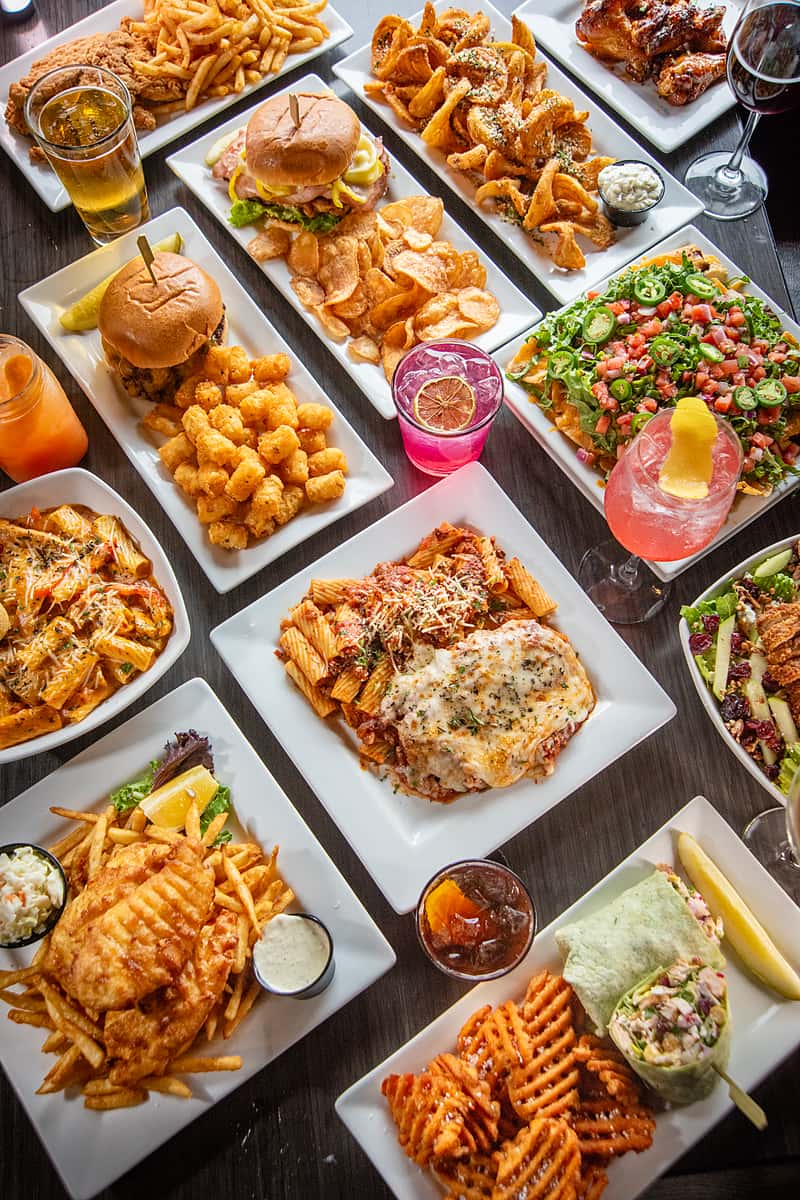 Top view of a restaurant table filled with a wide variety of American dishes including burgers, fries, pasta, nachos, wings, wraps, and colorful drinks
