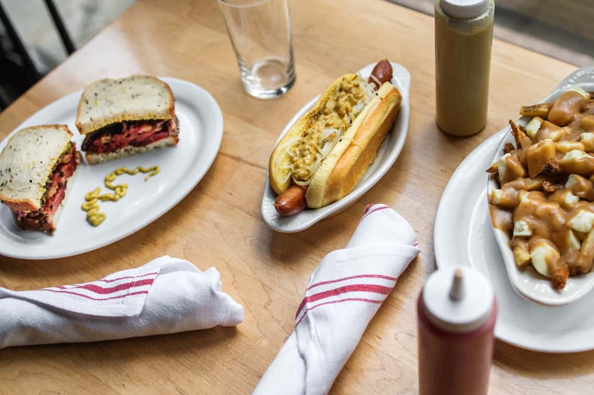 A pastrami sandwich with mustard, a hot dog topped with onions and mustard, and poutine with gravy and cheese curds.