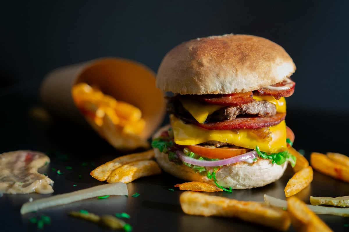 A fully-loaded burger with toppings and fries are displayed on a dark surface from Spot Gourmet Burgers.