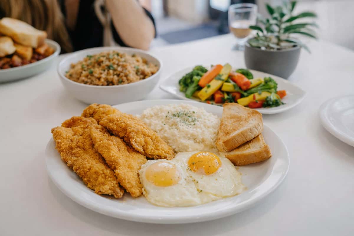 Plate with fried fish fillets, two sunny-side-up eggs, rice, toast, and mixed vegetables.