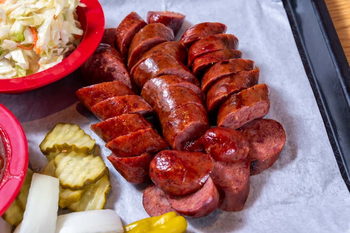 Sliced smoked sausage with a glossy BBQ finish served alongside coleslaw, pickles, and onions on a Southern barbecue tray.
