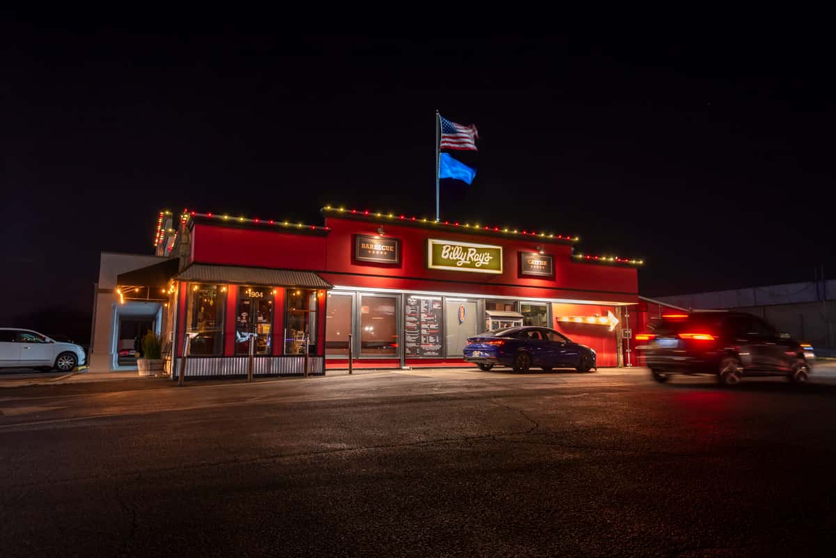 Exterior of a Southern barbecue restaurant at night with a bright red building, illuminated signage, string lights, and cars parked outside.
