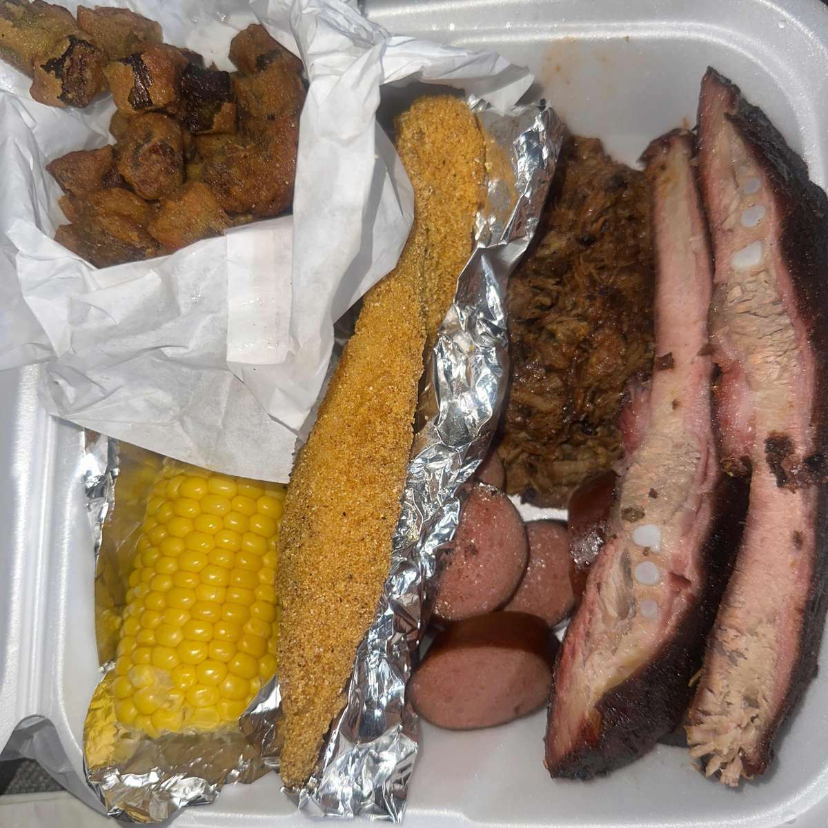 Loaded barbecue platter featuring fried okra, corn on the cob, fried catfish, sausage, pulled pork, and smoked ribs — a variety-filled Southern comfort feast in a takeout box.