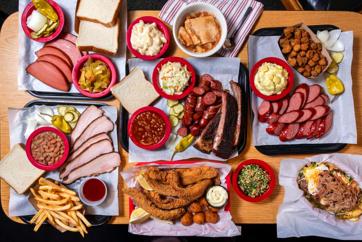 Southern barbecue spread with smoked brisket, sliced sausage, pulled pork, fried chicken tenders, baked beans, coleslaw, potato salad, white bread, pickles, and classic sides on a rustic table.
