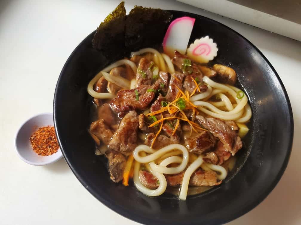 Classic Japanese noodle soup dish featuring thick, chewy udon noodles in a savory and fragrant broth made with soy sauce, mirin, and dashi, topped with tender slices of beef, green onions, and fish cake