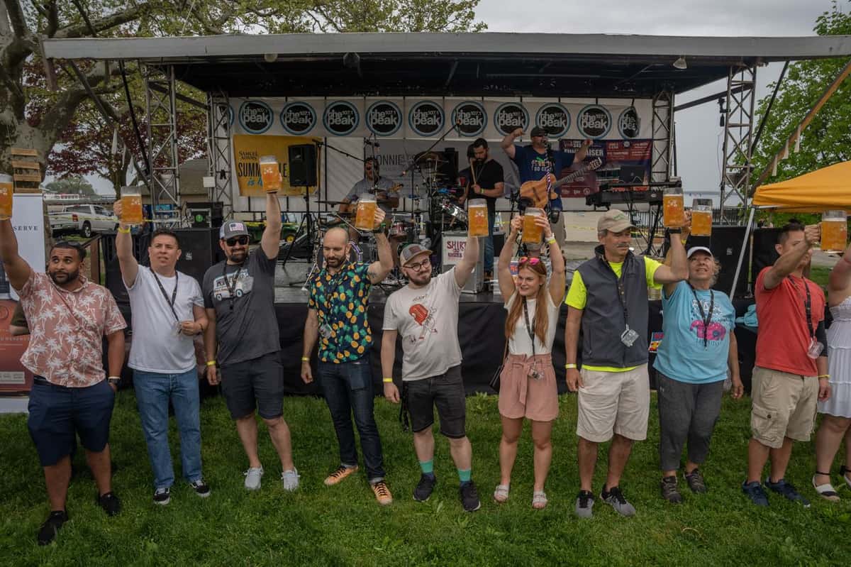 Contestants stand in a row holding beer steins aloft in front of a stage with a live band at an outdoor beer festival.