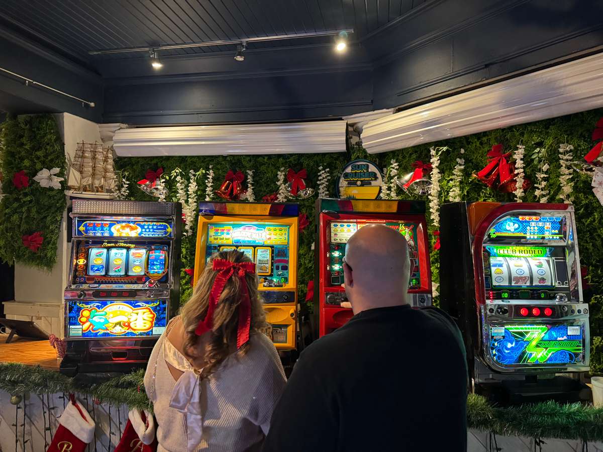 Man and woman playing colorful slot machines in a festively decorated room with garlands, red bows, and Christmas stockings.