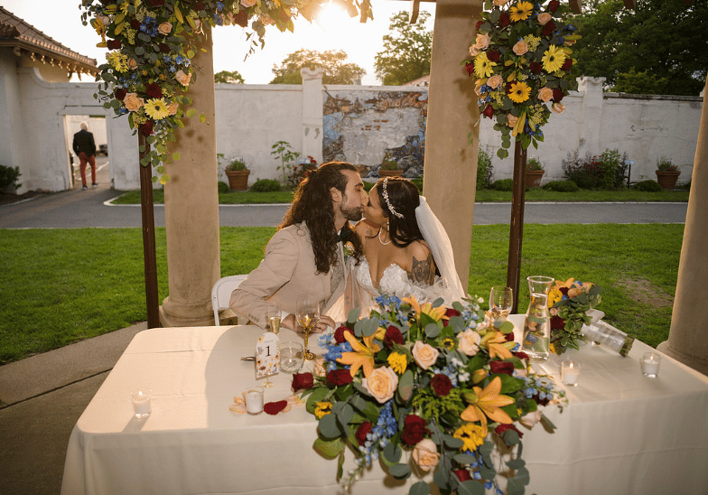 Bride and groom share a kiss at their outdoor wedding reception, seated at a flower-adorned table under a decorated archway.