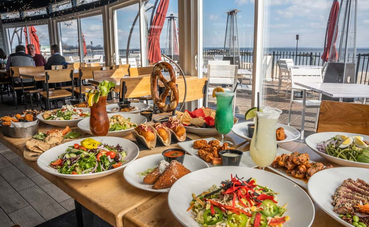 Assorted appetizers, salads, tacos, and cocktails spread across a large table in a beachside restaurant with an ocean view.