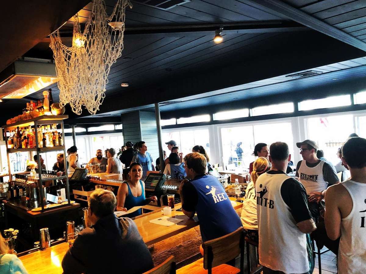 Casual bar scene with groups of people in sports jerseys socializing and enjoying drinks around a wooden bar in a bright, lively space.