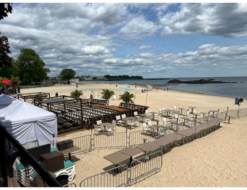 Beachfront setup with a wooden deck, lounge chairs, and tables enclosed by fencing, overlooking the ocean under a partly cloudy sky.