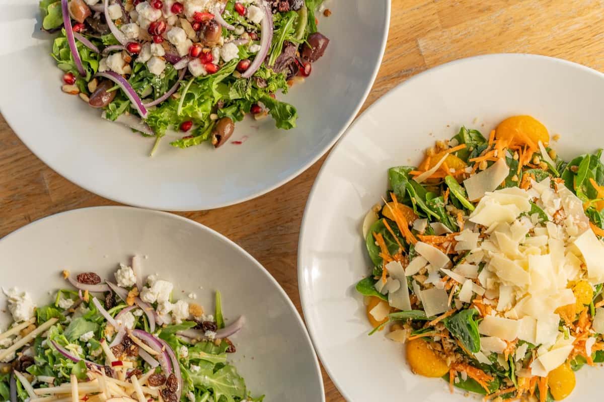 Three fresh salads in white bowls with colorful ingredients like shaved cheese, pomegranate seeds, olives, and mixed greens.