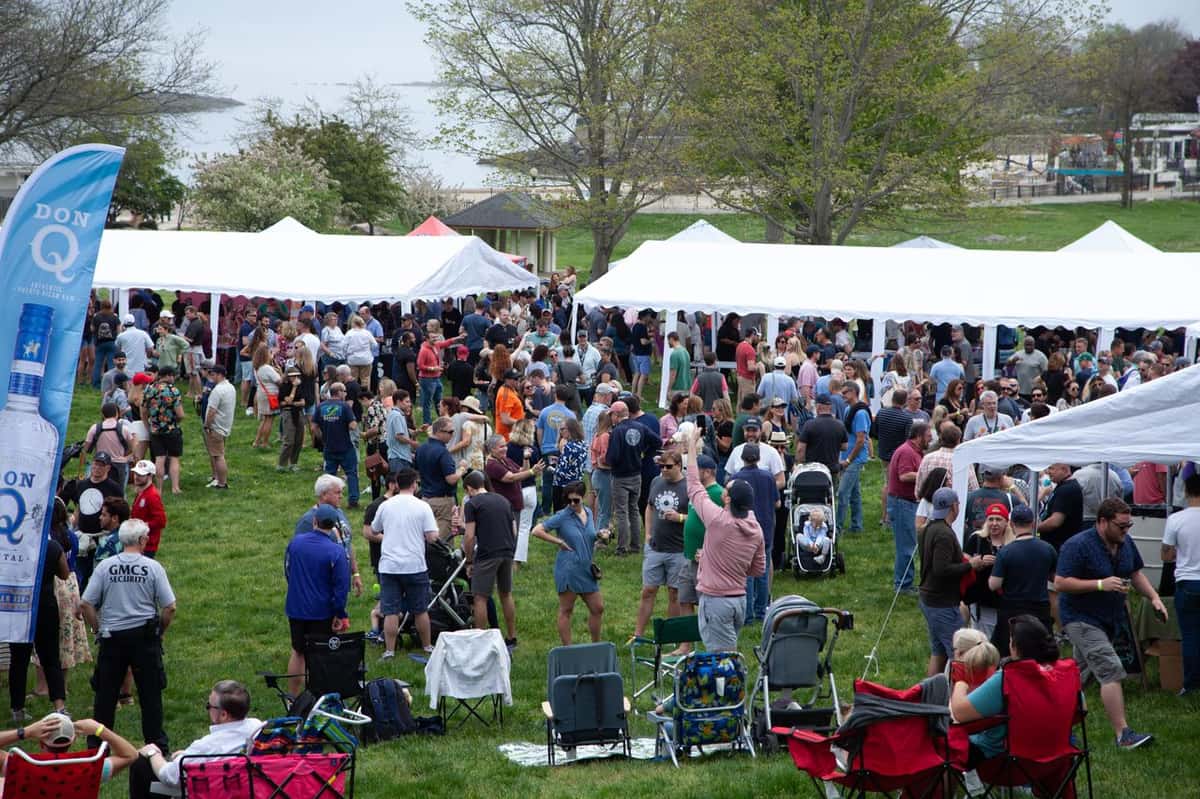 Large crowd gathers on a grassy lawn under white tents for a daytime outdoor festival near the waterfront.