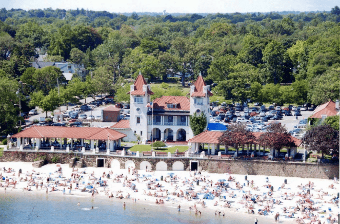 Aerial view of a crowded beach in front of a historic, castle-like building with red rooftops, surrounded by trees and a full parking lot.