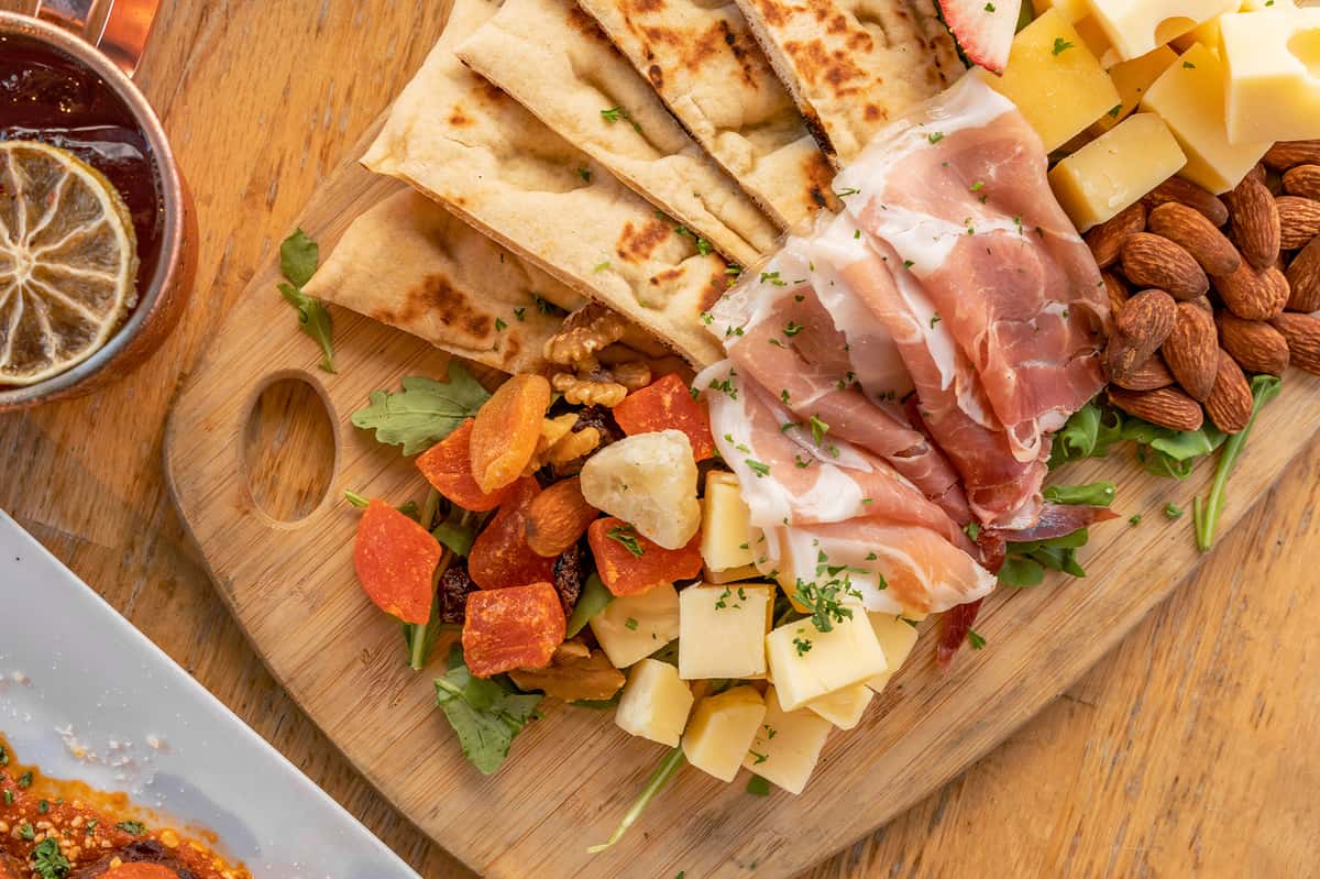 Charcuterie board with sliced flatbread, prosciutto, cubed cheeses, almonds, dried fruits, walnuts, and fresh greens.