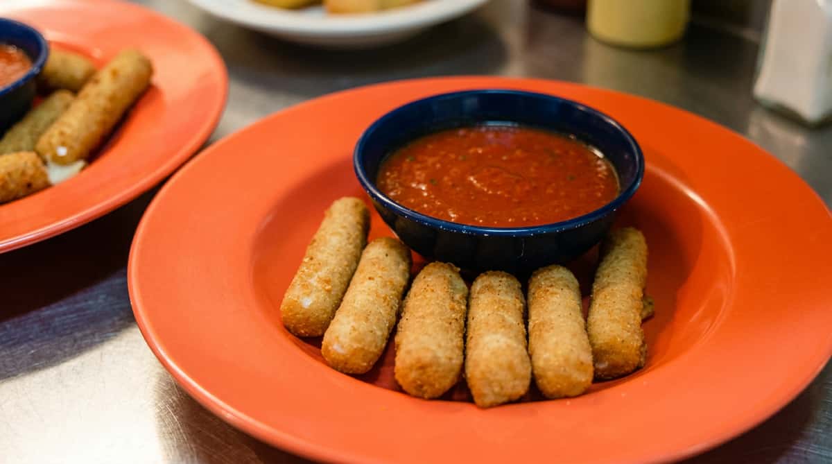 Mozzarella sticks on an orange plate with a dark blue bowl of marinara sauce.