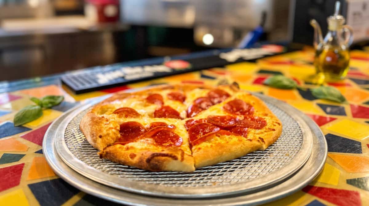 Pepperoni pizza on a metal pan, with a mosaic pizza oven and colorful tabletop blurred in the background.