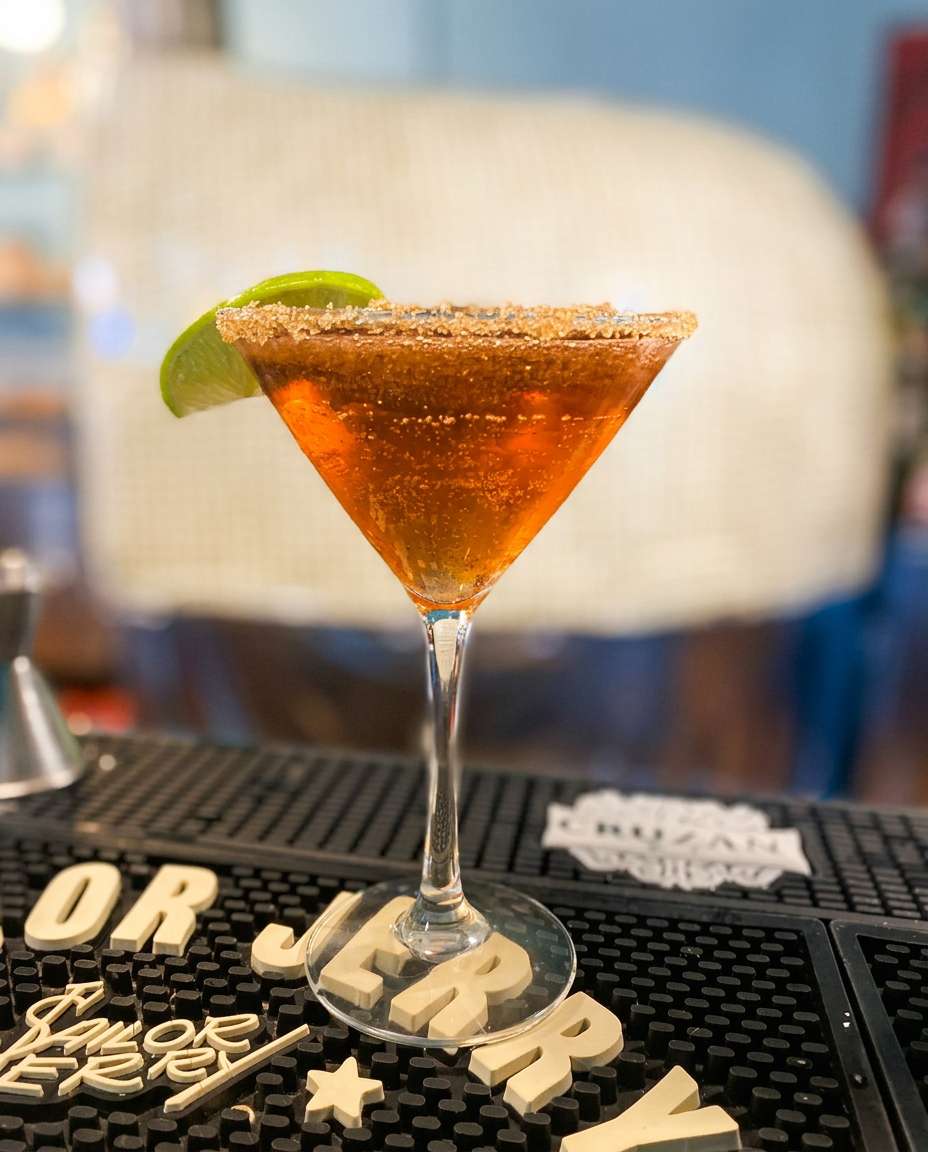 Martini glass with a brown-orange drink, sugar-rimmed, and a green lime slice, on a black bar mat.
