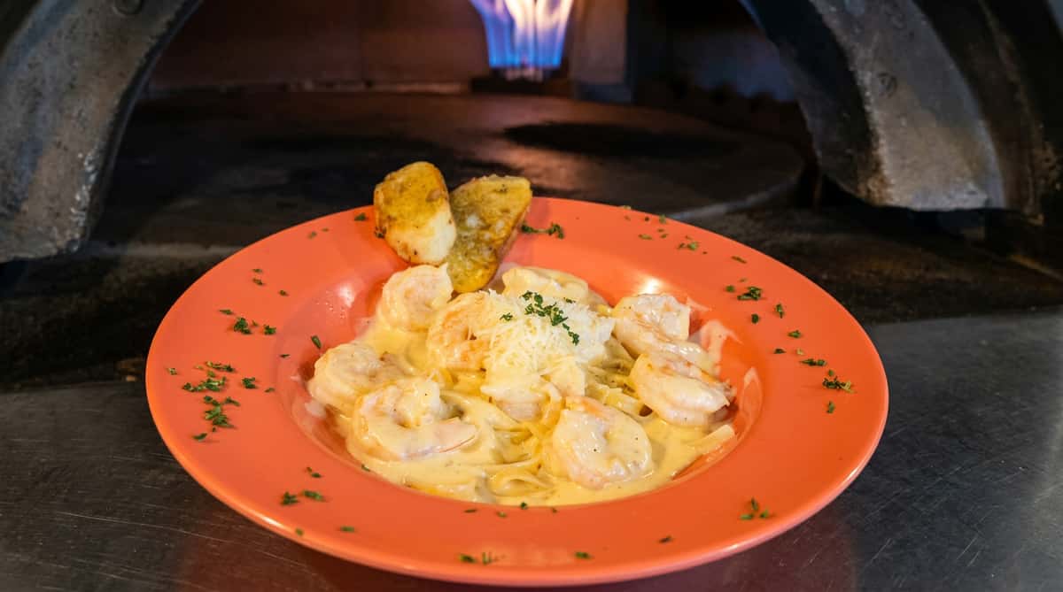 Shrimp alfredo pasta with garlic bread on an orange plate, in front of a wood-fired oven with blue flames.