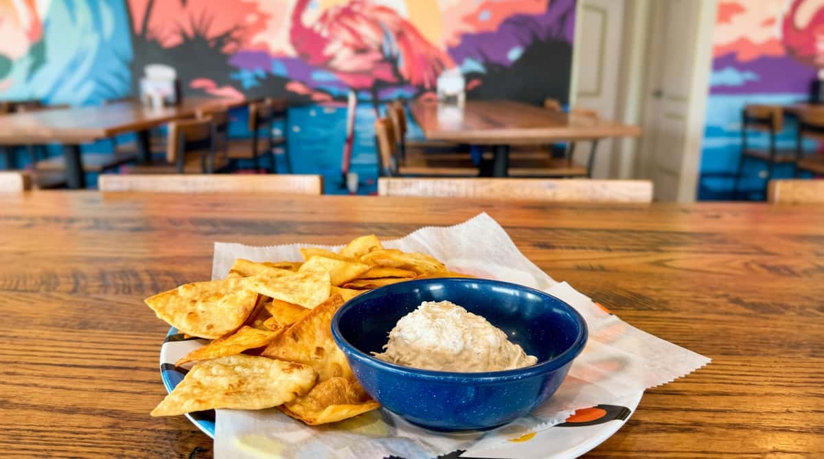 Fried tortilla chips and a blue bowl of white dip on a plate, with a colorful flamingo mural in the background.