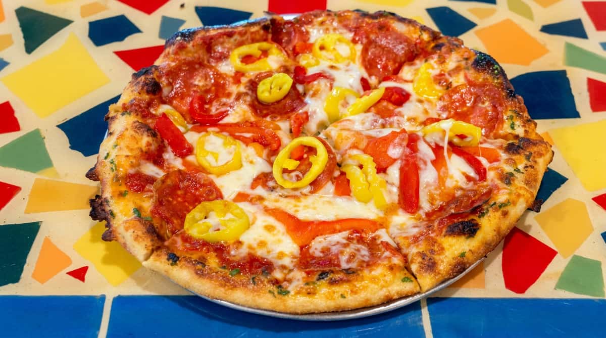 Pizza with pepperoni, red bell peppers, banana pepper rings, and melted cheese on a colorful tiled surface.
