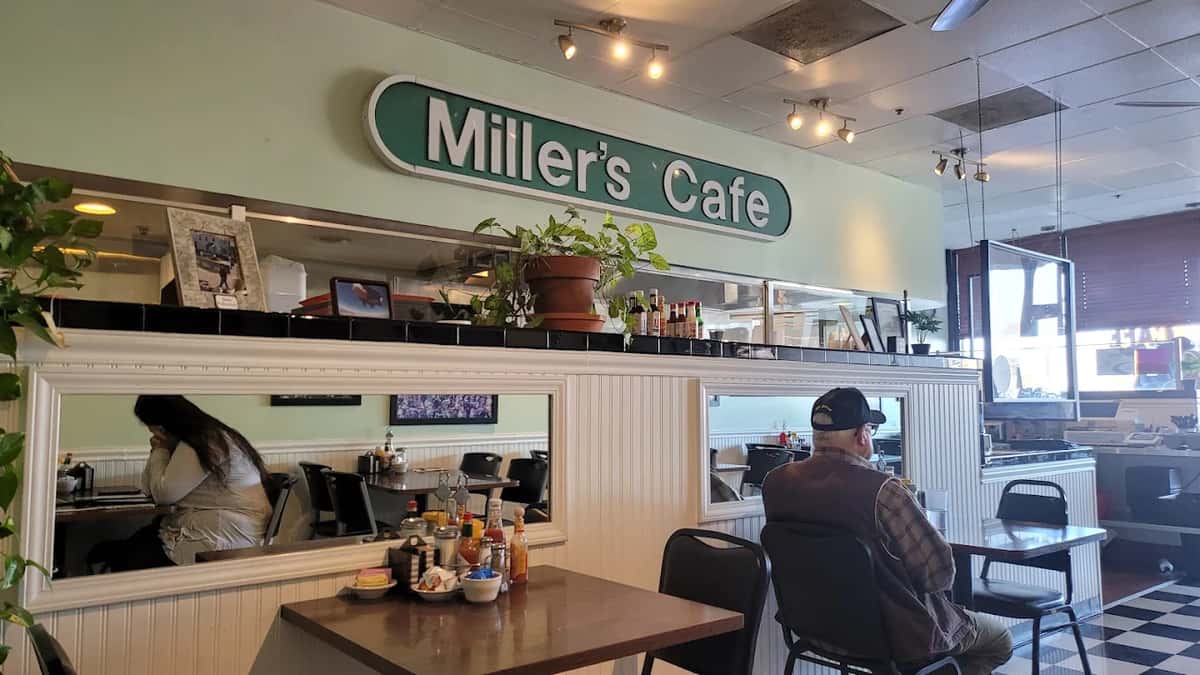 Interior of Miller's Cafe diner showing the green sign, counter pass-through, and patrons seated at tables near the black and white checkerboard floor.