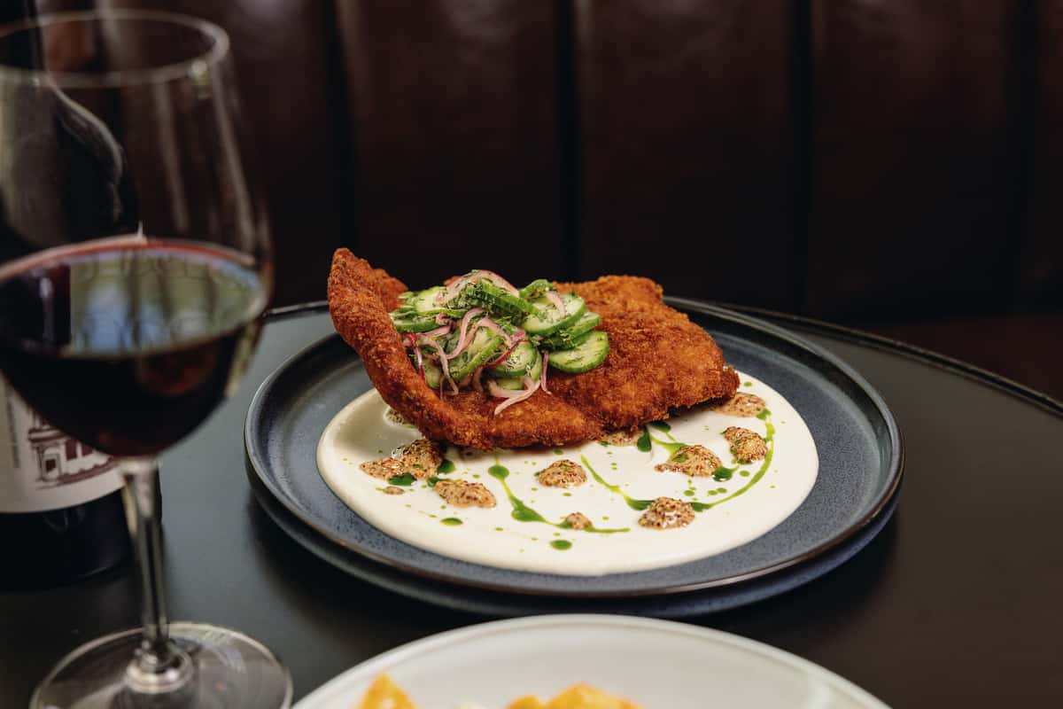 A beautifully plated chicken dish on a round black plate, served with a creamy sauce and garnished with cucumber and herbs.