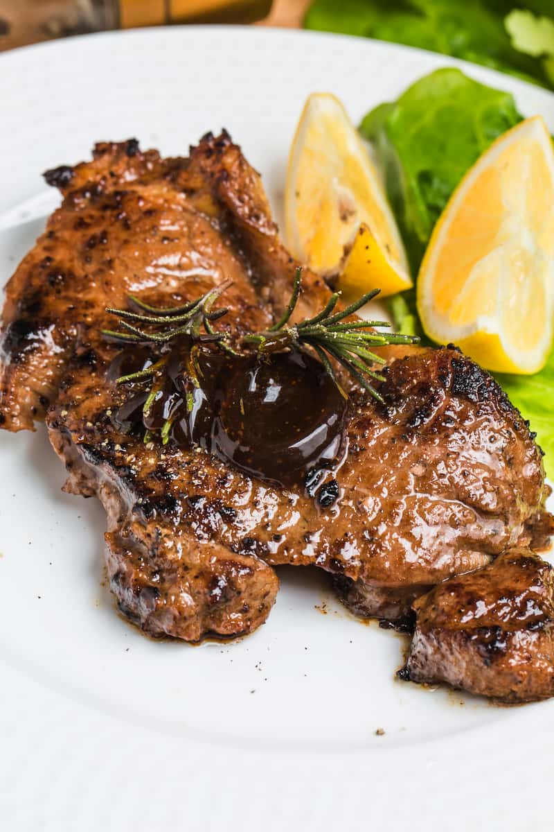 Grilled steak with a glossy sauce and rosemary sprig, served with lemon wedges and romaine lettuce on a white plate.