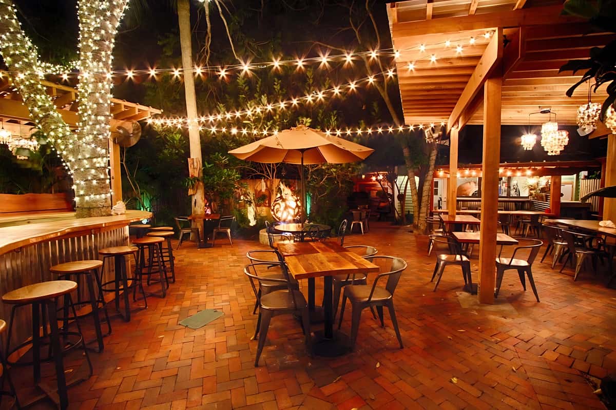 Outdoor garden dining illuminated by string lights and chandeliers, featuring brick flooring and cozy seating areas.