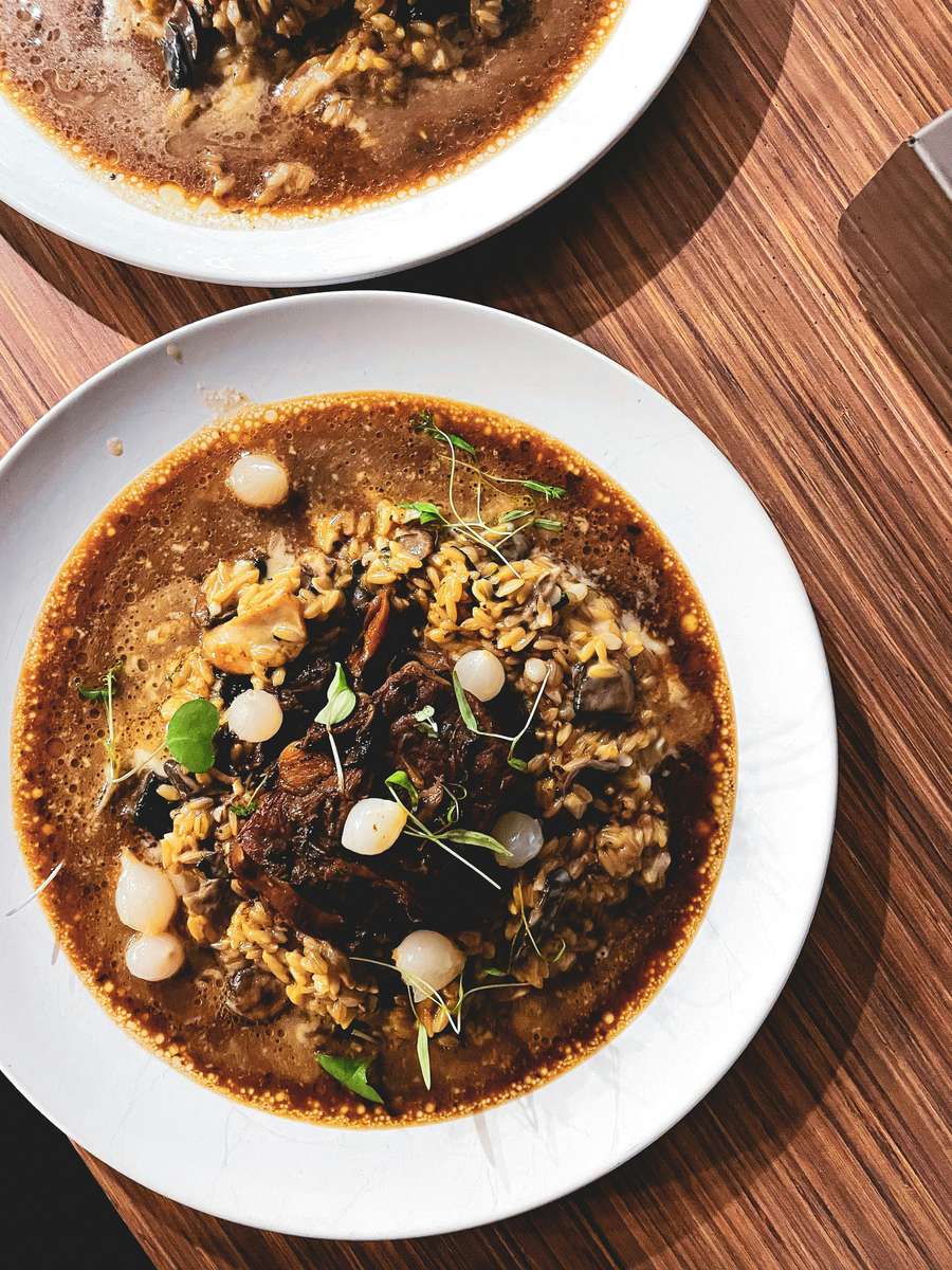 Deep, flavorful oxtail stew served over risotto-style rice, garnished with pearl onions and microgreens, in a shallow white bowl on a wooden table.