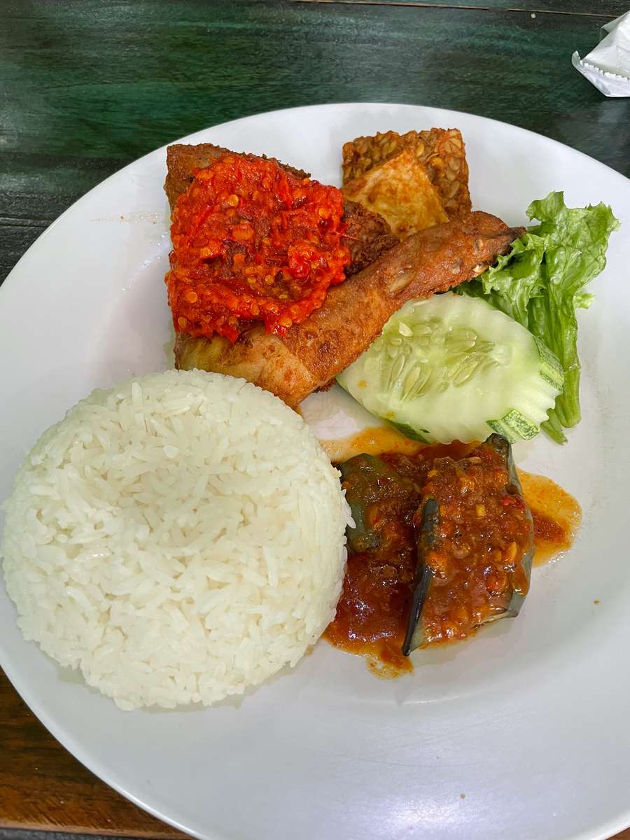 Plate of Indonesian nasi ayam geprek with steamed white rice, crispy fried chicken topped with sambal chili paste, fried tempeh, fresh cucumber, lettuce, and a side of spicy eggplant in chili sauce.